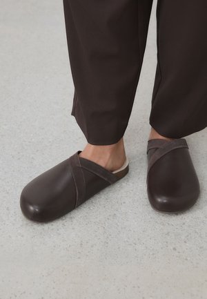 Stopy w ciemnobrązowych wsuwanych butach z szerokimi zaokrąglonymi noskami i miękkimi paskami, połączone z ciemnobrązowymi spodniami, stojące na jasnej, cętkowanej podłodze.