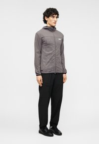 Γκρι ζακέτα με φερμουάρ μπροστά και κουκούλα, με λεπτό μοτίβο και ελαστικές μανσέτες, συνδυασμένη με μαύρο φαρδύ παντελόνι και μαύρα παπούτσια.