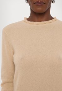 Gros plan d'une personne portant un pull beige en maille avec un col à volants, montrant les épaules et les lèvres sur un fond blanc.