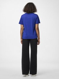 Person med lockigt hår står vänd bort från kameran och bär en kungligt blå t-shirt, vida svarta byxor och vita skor med ränder mot en enfärgad bakgrund.