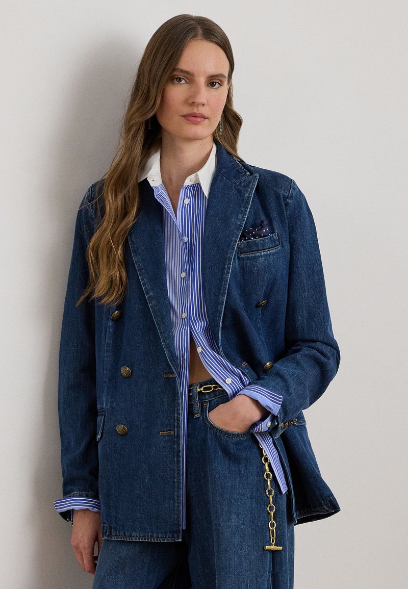Kvinde med langt bølget hår iført en blåstribet skjorte, dobbeltradet denimblazer med guldknapper og matchende denimbukser med kædebælte.