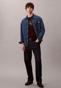 Camisa de mezclilla con cierre de botones, azul oscuro; camiseta burdeos con logo estilizado; pantalones oscuros; zapatos negros; fondo neutro.