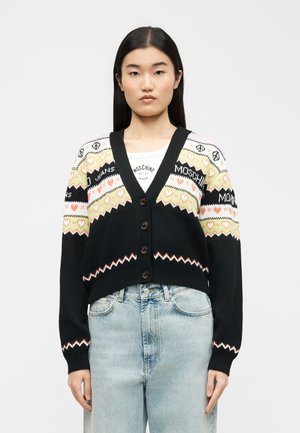 Tânără purtând un cardigan negru cu nasturi, decorat cu modele de inimi galbene și roz, asortat cu blugi albaștri deschiși, cu talie înaltă.