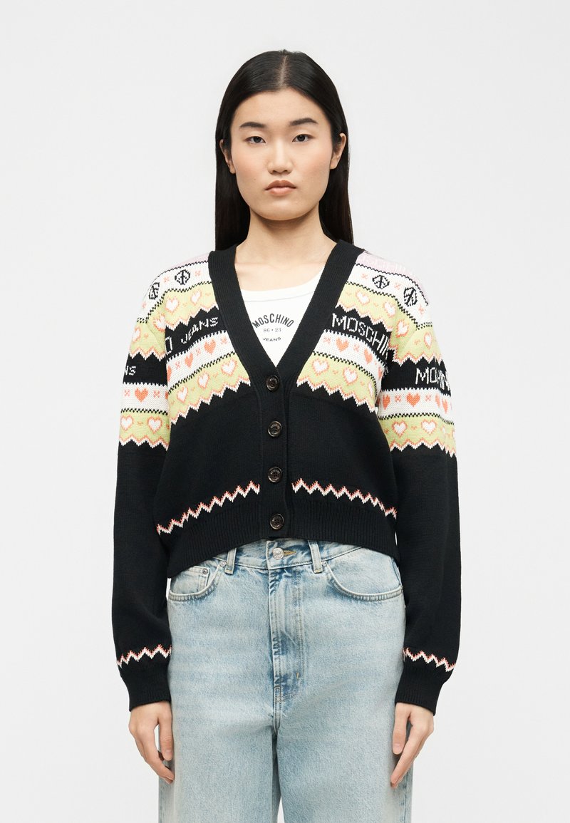 Νεαρή γυναίκα που φοράει μαύρη ζακέτα με κουμπιά και σχέδια από κίτρινες και ροζ καρδιές, συνδυασμένη με ανοιχτόχρωμα τζιν ψηλόμεσα.