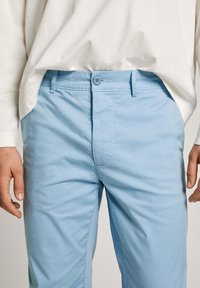 Gaiši zili chinos ar gludu tekstūru, ar pogu aizdari un jostas cilpām. Apvienoti ar baltu krekli ar garām piedurknēm.