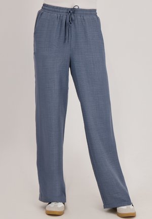 Blauwe losse broek met elastische tailleband en trekkoord, gedragen met witte sneakers op een lichte achtergrond.