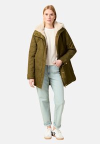 Parka verde olivo con capucha de pelo, cierre de cremallera y bolsillos, combinada con jeans de color azul claro y zapatillas blancas.