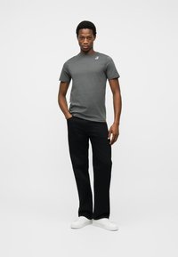 Νέος άνδρας όρθιος, φοράει γκρι κοντομάνικο μπλουζάκι, μαύρο παντελόνι, λευκά αθλητικά παπούτσια, με το ένα χέρι στην τσέπη, μπροστά σε λευκό φόντο.