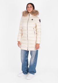 Crème pufferjas met een bontgevoerd capuchon, ritssluiting aan de voorkant en twee zakken aan de zijkant. Draag het over lichtblauwe wide-leg jeans en witte sneakers.