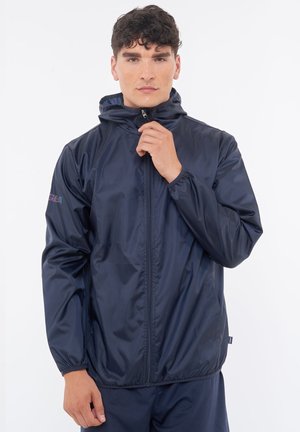 Junger Mann trägt eine marineblaue Windjacke mit Reißverschluss und Kapuze, steht vor einem schlichten weißen Hintergrund.