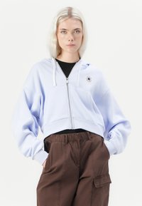 Sudadera corta de color azul claro con cremallera en la parte frontal y cordones blancucos, que presenta un parche con un logo. Combinada con pantalones cargo marrones con bolsillos.