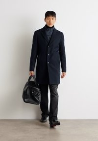 Casaco de lã azul-marinho com um corte ajustado, dois botões e bolsos laterais. Usado sobre uma jaqueta preta de gola alta, combinado com calças e sapatos pretos.