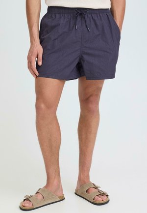 Mann trägt marineblaue Shorts mit Kordelzug und beigen Wildleder-Sandalen mit Doppelverschluss, steht vor einfachem Hintergrund.