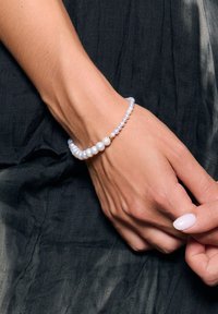 Bransoletka z białymi perłami i małymi niebieskimi koralikami, akcentowana złotym detalem. Noszona na dłoni na ciemnym, teksturowanym tle.