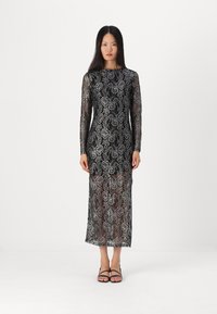 Robe en dentelle noire à manches longues avec des motifs floraux complexes, un corsage ajusté et une jupe semi-transparente. Des sandales noires complètent le look.