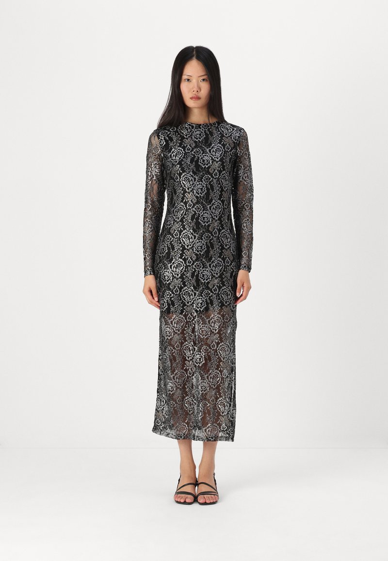 Robe en dentelle noire à manches longues avec des motifs floraux complexes, un corsage ajusté et une jupe semi-transparente. Des sandales noires complètent le look.