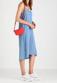 Džínové midi šaty ve světle modré barvě s výstřihem do V, doplněné malou červenou přes shoulder taškou a bílými teniskami, s uvolněným střihem.