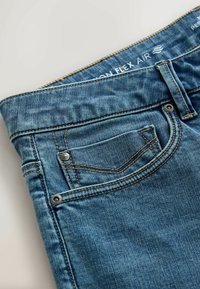 Zone de la taille d’un jean en denim bleu montrant la poche avant, petite poche à monnaie avec rivets métalliques, passants pour ceinture et texte « ON FLEX AIR » à l’intérieur de la ceinture.