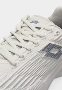 Makro záber bielej športovej topánky s textúrovaným povrchom, bielymi šnúrkami a sivým geometrickým logom na boku.