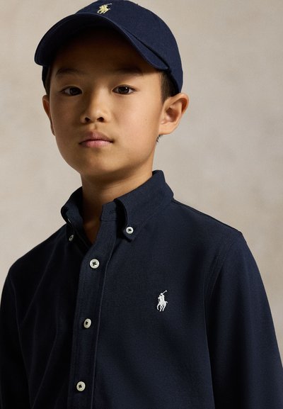 Jaunas berniukas dėvi tamsiai mėlyną marškinių tipo palaidinę mygtukinę su derančia kepure, ant abiejų yra mažas baltas polo žaidėjo logotipas.