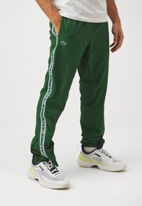 Pantaloni della tuta verdi con una striscia bianca laterale, polsini elasticizzati e logo Lacoste. Indossati con sneakers bianche e grigie con un accento neon.