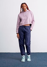 Haut de survêtement court violet clair avec capuche, associé à un jogging à taille élastique bleu marine et des baskets montantes bleu clair et blanches.