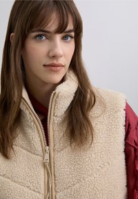 Chaleco de forro polar beige con tejido texturizado, cierre de cremallera y cuello alto; superpuesto sobre una camisa roja con mangas con volantes.