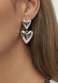 Orecchini pendenti d'argento a forma di doppio cuore indossati in un primo piano di un orecchio femminile con capelli castano chiaro visibili.