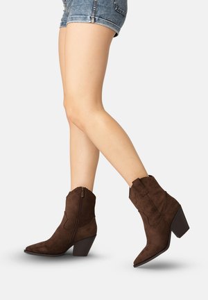 Piernas con pantalones cortos de mezclilla y botines de ante marrón con cremallera lateral y tacones cuadrados, un pie ligeramente levantado.