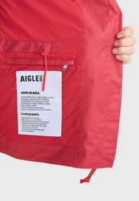 Rote wasserdichte Jacke mit einer Innentasche, die ein weißes Etikett mit Text enthält. Glatte Textur mit einem Zugband am Saum.
