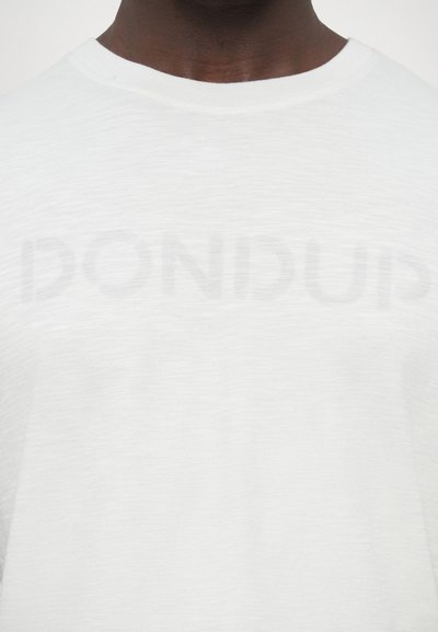 T-shirt blanc à col rond en tissu léger avec une texture subtile ; présente un logo délavé en grosses lettres capitales sur la poitrine.
