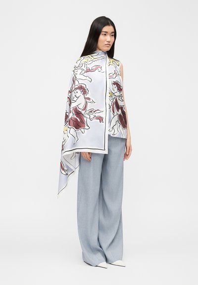 Γυναίκα με μακριά σκούρα μαλλιά που φοράει φαρδιά ανοιχτό μπλε παντελόνια, λευκά μυτερά παπούτσια και ένα μεγάλο λευκό φουλάρι με κόκκινα και κίτρινα σχέδια αγγέλων.