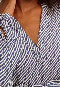 Blåt og hvidt mønstret stof med lodrette striber og geometriske motiver, med V-udskæring og løs pasform. Glat tekstur.