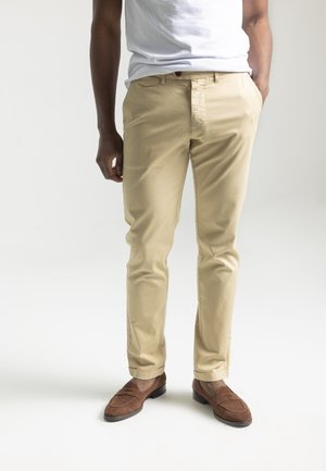 Mann trägt beige Chinos mit Händen in den Taschen und braunen Wildleder-Loafers, steht vor einem schlichten hellen Hintergrund.