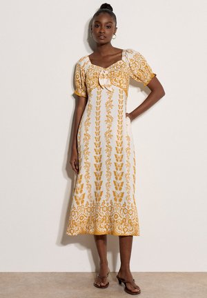 Femme debout portant une robe midi blanche avec des motifs floraux et papillons jaune moutarde, des manches bouffantes et un détail noué à l'avant.