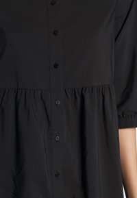 Camicia nera con bottoni a pressione, caratterizzata da una vita arricciata, maniche corte e una superficie in tessuto morbido con bottoni simili a perle.