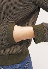 Szara sweterka z siateczki z żebrowanymi zielonymi mankietami. Posiada boczną kieszeń zintegrowaną z designem, podkreślającą oddychalność i teksturowany materiał.