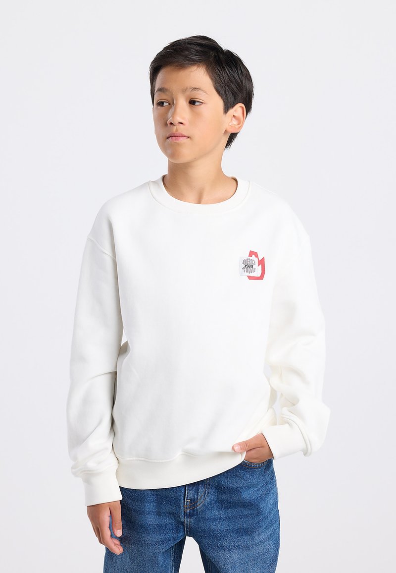 Valkoinen collegepaita pyöreällä kaula-aukolla ja pitkillä hihoilla, jossa on pieni punainen ja harmaa grafiikka rinnan kohdalla. Pehmeä, tekstuurinen kangas.