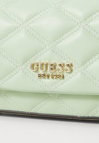 Jasnozielona torebka z faux skórki z pikowanym wzorem, z złotym logo "GUESS" i akcentami w szwach. Kompaktowy prostokątny kształt.