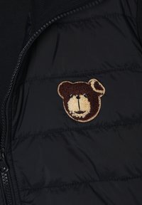 Sort polstret vest med lynlås, der har et struktureret brunt og cremefarvet teddy-bjørn-patch på brystområdet. Glat stof med quiltet design.
