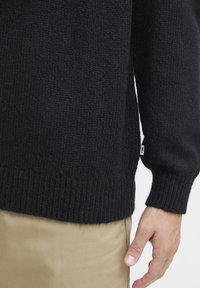 Suéter negro de punto con puños y dobladillo acanalados, que presenta un ajuste holgado y una tela texturizada, llevado sobre pantalones de color claro.