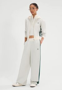 Jasnoszary zestaw sportowy, w tym bluza z kapturem na zamek z kremowymi akcentami oraz spodnie z szerokimi nogawkami z zielonymi paskami po bokach. Gładka tekstura i luźny krój.