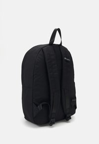 Sac à dos en tissu noir avec une forme ergonomique, des bretelles rembourrées et une poignée supérieure. Présente une marque minimale d'un côté et pas de motifs visibles.