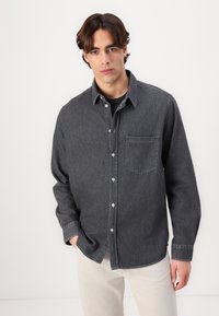Graues Denim-Hemd mit Knopfleiste, langen Ärmeln, einer Brusttasche und weichem Material. Über einem schwarzen T-Shirt getragen und kombiniert mit hellen Hosen.