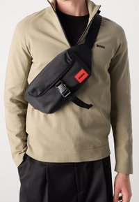 Man som bär en beige zip-up tröja och svarta byxor, med en svart axelremsväska som har en röd "HUGO"-etikett och plastspänne.