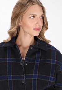 Blond kvinna med vågigt hår iklädd en mörkblå rutad jacka med stora knappar, tittar åt höger mot en vit bakgrund.
