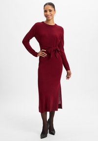 Strickmidi-Kleid in Burgunderrot mit langen Ärmeln, einem Taillengürtel und seitlichem Schlitzdetail. Getragen mit schwarzen spitzen Absätzen. Glatte Textur.