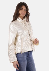 Giacca corta color crema, lucida, con colletto alto, zip frontale e tasche laterali; design minimalista e texture liscia.