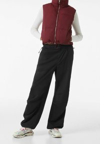 Burgundowy kamizelka z ociepleniem, z wysokim kołnierzem, suwakowym przodem i ściągaczem u dołu, zestawiona z czarnymi spodniami o szerokich nogawkach i wielokolorowym obuwiem sportowym.