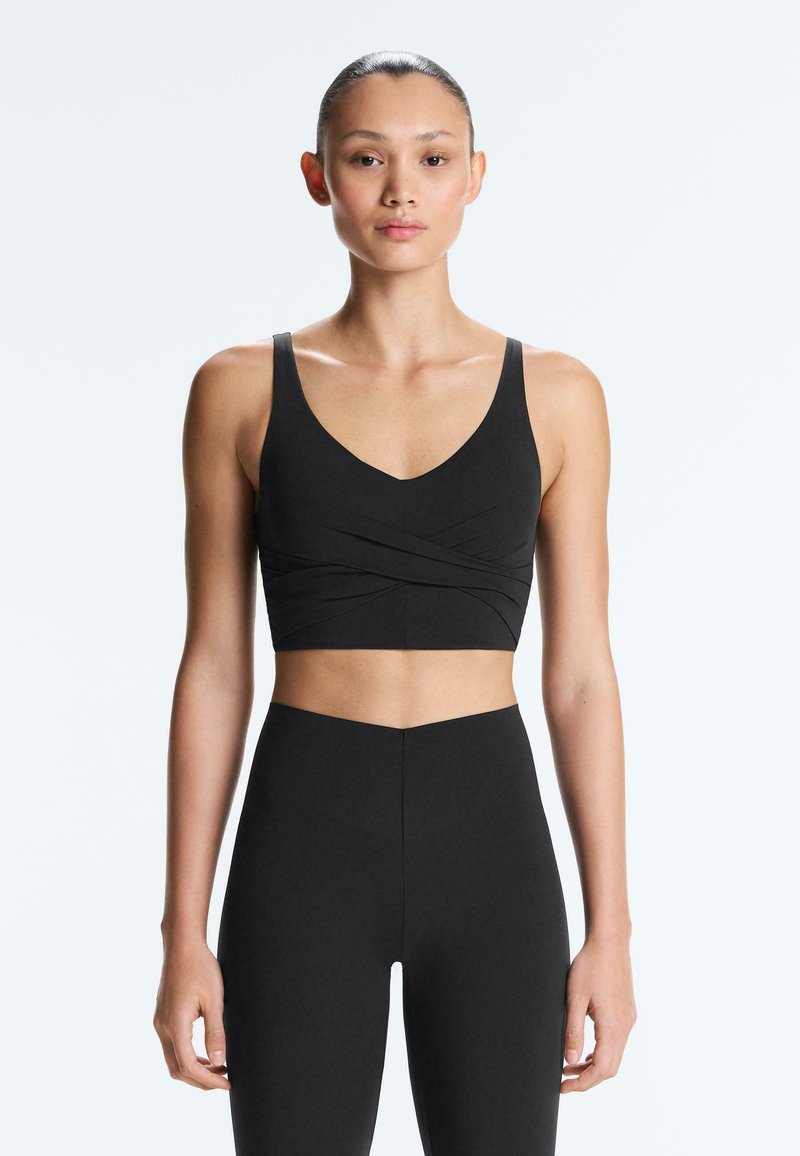 Žena čelem k nám, oblečená v černé sportovní podprsence s překříženými detaily a černých legínách s vysokým pasem na bílém pozadí.